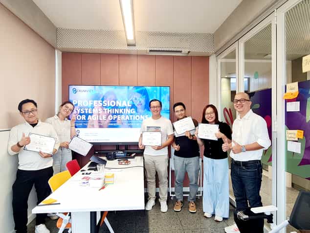 Học viên cầm chứng nhận chụp ảnh cùng trainer trước màn hình Professional Systems Thinking for Agile Operations