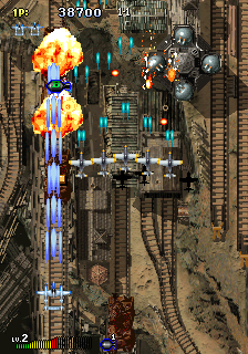 A vertical scrolling shooter displays a military-themed level with industrial structures, railroad tracks, and fortifications rendered in isometric perspective. The player's aircraft occupies the lower center, firing upward as orange explosions erupt across the screen from weapon fire and enemy contact. Blue enemy bullets descend toward the player position. The HUD shows a score of 38700 at top left with remaining lives indicated. Enemy aircraft and ground installations are visible throughout the upper and middle sections. The color palette emphasizes grays, browns, and metallic tones with bright orange and blue accents for explosions and projectiles.