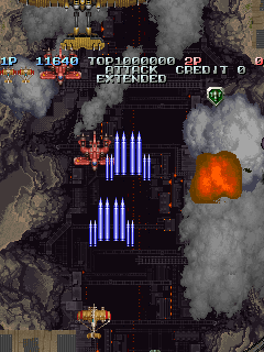 A vertical scrolling shoot-em-up displaying a top-down view of a military base under aerial attack. Blue vertical columns of enemy fire rise from the center-lower portion of the screen, while orange explosions burst across the terrain. The HUD shows score, lives remaining, and weapon credit at the top-left in yellow text. Pixelated sprites of aircraft and ground structures are visible against a dark, smoky battlefield with brown and gray tones indicating destroyed installations. The image captures mid-action gameplay with active combat exchanges between the player and multiple on-screen threats.