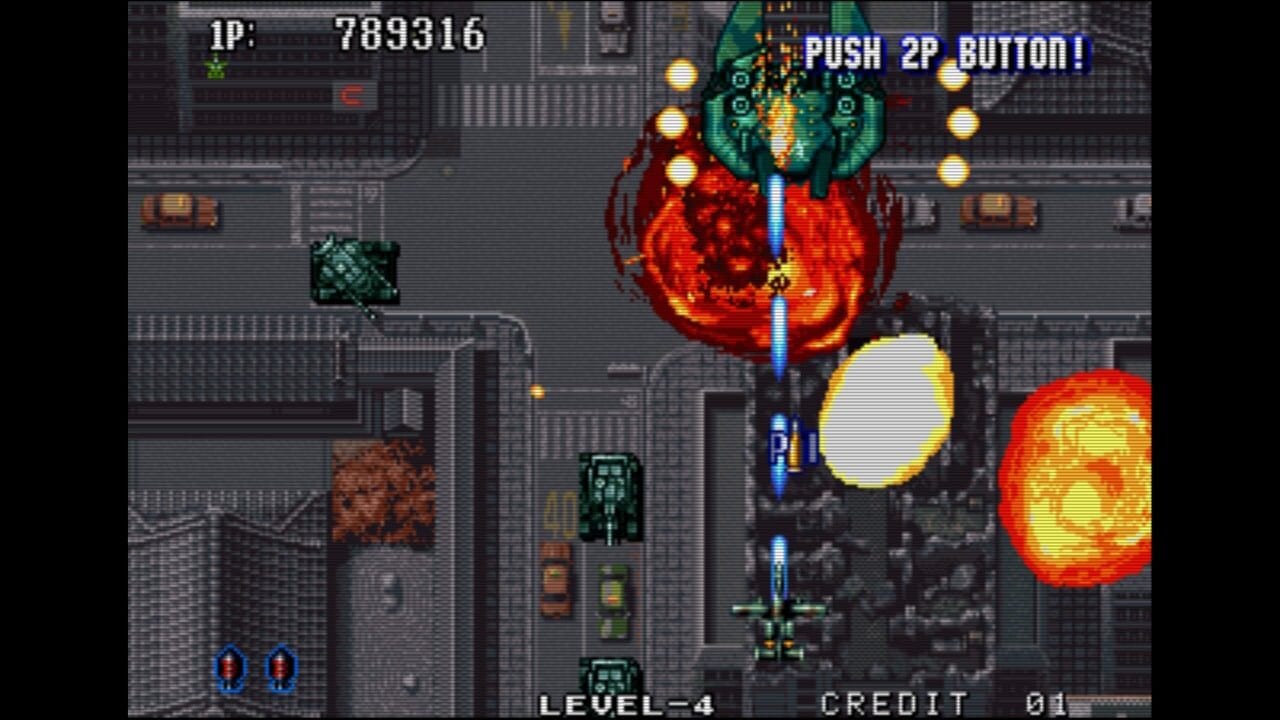 A top-down vertical shooter displays a green player aircraft centered in the lower portion of the screen, firing upward at enemies. A large red and orange flame-engulfed boss structure dominates the upper-center area, with bright yellow orbs and smaller enemy sprites scattered around it. The background shows an industrial cityscape with gray buildings and brown rubble. Score displays 789316 in the top-left corner, with "PUSH 2P BUTTON!" text in the top-right. Level-4 and Credit 01 appear in the bottom corners. Yellow and orange circular projectiles fill the screen.