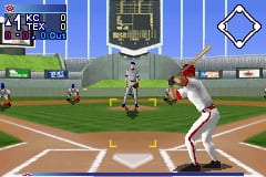 A batter in red stands at home plate in a batting stance, facing a pitcher on the mound. The baseball field displays green grass, brown dirt infield, and a gray outfield wall. The HUD shows a small score panel in the upper left corner with game information, while a diamond-shaped icon appears in the upper right. Multiple fielders are positioned across the field in standard defensive formation. The sprites use low-resolution pixel graphics typical of Game Boy Advance sports games, with a bright blue sky occupying the upper portion of the screen.