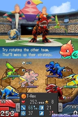 A battle screen displays two teams of fossil fighters in a stadium arena with crowded bleachers and a large inflatable mascot above the dugout. The player's team occupies the lower half with four creatures arranged in a row, including a green insectoid, a beige quadruped, a blue bipedal fighter, and a red creature. The opponent's team is positioned higher on the field with a brown beast-like creature prominently featured. A scrolling text message reads "Try rotating the other team. That'll mess up their strategy." The bottom UI panel shows character stats, health bars, and numerical values including "T-Rex," "253," "37," and "FP" indicators. The scene uses a colorful sprite-based art style with pixelated graphics typical of Nintendo DS hardware.