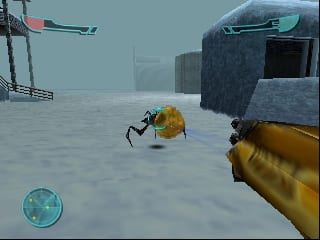 A first-person perspective shows a yellow armored soldier's arm holding a weapon in the lower right. An enemy figure in yellow armor stands ahead in a snowy outdoor environment with industrial structures and buildings visible in the hazy background. A radar or minimap display appears in the lower left corner. The scene uses a muted blue-gray color palette typical of N64-era graphics with visible polygon-based geometry.