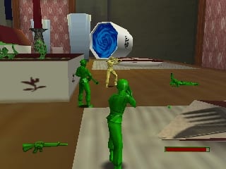 A green plastic soldier action figure stands in the center of an indoor room holding a large blue and white spherical object above its head. The soldier is rendered in low-polygon 3D graphics typical of N64 hardware. The room features brown wood flooring, pink and red striped wallpaper, a desk with a white monitor and potted plant on the left side, and scattered green toy soldier figures and weapons on the ground. A health bar appears in the lower right corner of the screen. The lighting casts shadows across the floor, and the overall scene depicts a miniaturized domestic interior environment.