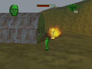 A green plastic soldier stands in the center of an outdoor dirt area with corrugated metal structures visible on the left and hillside terrain on the right. A yellow explosion effect occurs near the soldier's position. The soldier's health bar appears at the top left in red and green, with the number 90 displayed in the top right corner. The low-polygon 3D environment uses a muted brown and green color palette typical of N64-era graphics.
