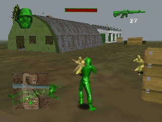 A green plastic soldier character stands in the center of an outdoor military compound. A large green barracks building with a gray roof dominates the left side, flanked by smaller structures and wooden crates scattered across tan dirt ground. A rifle with ammunition counter displays in the upper right. The HUD shows health status and weapon inventory in the lower left corner. Two enemy soldiers appear in the distance on the right. The 3D environment uses muted earth tones with low-polygon geometry typical of late-1990s console graphics.