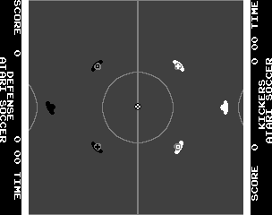 A split-screen soccer field viewed from above shows two halves separated by a center line with a circle marking the midfield. A white diamond-shaped marker appears at the center. Small white and dark player sprites are scattered across both field halves, positioned at various locations. The right side displays score information and game text in small white lettering along the edges. The overall visual style uses a dark gray background with white vector-style graphics typical of early 1980s arcade games.