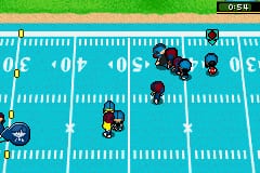 A football field viewed from above shows two teams of pixel-art players positioned across the turquoise grass with white yard markings. Multiple dark-colored players cluster in the center-left area, while lighter-colored players are scattered across the field. The scoreboard displays 0-0 at the top left, and a small icon in the upper right corner indicates the current game mode. The 16-bit sprite graphics and simplified player models are typical of Game Boy Advance sports games.