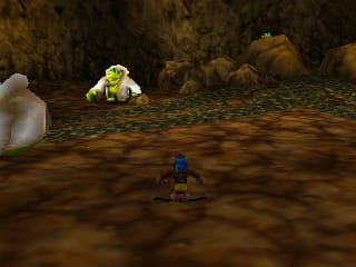 A small blue character stands in the center of a brown dirt ground area surrounded by rocky cave walls. A large yellow-green creature with a round body sits on the left side of the scene, while a white ethereal figure appears on the far left. The environment uses a brown and tan color palette with rough stone textures typical of N64-era 3D rendering. The perspective is from a third-person camera angle positioned behind the player character.