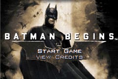 A Batman Begins title screen featuring a large close-up portrait of Batman's cowled face against a textured, weathered brown and tan background. The character's eyes glow white, and his facial features are rendered in a realistic style. The title "BATMAN BEGINS" appears in white capital letters at the top center. Below the title, two menu options are displayed in white text: "START GAME" and "VIEW CREDITS" in a sans-serif font. The overall color palette consists of muted earth tones with heavy shadows, creating a dark, atmospheric mood typical of Game Boy Advance graphics.