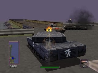 A tank with a panda symbol on its turret occupies the center foreground, colored dark blue with visible damage marks. Smoke plumes rise from explosions in the mid-ground and distance. To the left, a yellow construction crane sits on the industrial terrain. A second tank moves through the right side of the scene. The HUD displays a green health bar at the bottom left, a minimap in the upper left corner, and weapon/ammo indicators in the lower right. The environment is an industrial area with flat, textured ground beneath an overcast sky.