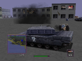 A large dark blue tank occupies the center of the screen, viewed from an isometric angle in a 3D rendered environment. A gray concrete building with multiple windows stands in the background. The lower-left corner displays a minimap with colored markers. A green health bar spans the bottom-center, with ammunition counter and other HUD elements visible on the right side. The ground is textured pavement with parked vehicles and industrial structures visible in the mid-distance.
