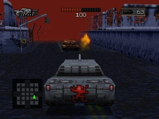 A gray tank with red accents occupies the lower center of the screen, viewed from above in a three-dimensional perspective. An explosion detonates in the middle distance across a dark water surface. A concrete platform spans the upper half of the frame, with red industrial structures and buildings visible along both sides. A HUD panel in the lower left displays a grid-based map with colored indicators. Numerical displays at the top show "100" and weapon/ammo counts. The overall color palette features muted reds, grays, and dark blues typical of N64-era graphics.