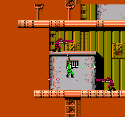 A top-down overhead view of a multi-level facility with orange exterior walls and brown-tan interior corridors. A green-suited bionic soldier stands in the center white chamber surrounded by yellow-green armored panels and machinery. Brown wooden platforms connect upper and lower sections. Red and brown enemy units are positioned throughout the environment. The 8-bit sprite artwork uses a limited NES color palette with detailed tile-based level design showing distinct interior and exterior areas across multiple vertical layers.