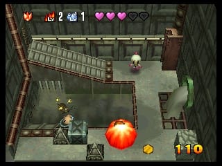 A top-down view of a tiled stone arena with brick walls forming a maze-like layout. A white character with a round head stands in the lower-left area. A large orange explosion effect with yellow sparks dominates the center of the screen. The upper-left UI shows a health bar with three red hearts, numbered counters displaying player statistics, and a bomb icon. The lower-right corner displays the number 110 in yellow text. The environment uses muted green and brown tones with 3D isometric-style graphics typical of early Nintendo 64 games.