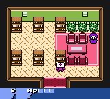 A top-down isometric perspective shows a interior room layout with brown wooden crates and barrels scattered across a tan floor, while the right side features a bright pink area with red furniture pieces. A small character sprite stands in the center of the room. The screen displays a blue status bar at the bottom with Japanese text and icons. The pixelated art style uses a limited color palette typical of Game Boy hardware, with the isometric view creating a pseudo-3D effect through angled tile placement.
