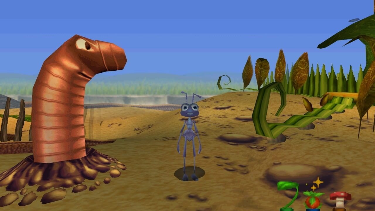 A blue ant character with large eyes stands in the center of a sandy desert landscape dotted with brown rocks. To the left, a large red worm-like creature emerges from the ground. Green plant-like structures and tall pointed vegetation fill the right side of the scene. A clear blue sky dominates the background with a distant treeline. Small colorful UI elements appear in the lower right corner, including green and red objects. The scene uses 3D polygonal graphics with a cartoony art style typical of early Nintendo 64 games.