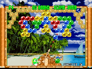 A puzzle game board displays colored spheres arranged in clusters against a tropical beach background with palm trees and ocean visible behind. The play field occupies the upper portion with various colored bubbles—red, blue, yellow, green—scattered across the grid. A wooden-framed border surrounds the play area. At the bottom, a cartoon character with an orange/brown appearance stands to the left side of the interface. The top of the screen shows green text displaying game information including score details. The art style uses bright, saturated colors typical of late-1990s arcade game sprites with 2D rendered backgrounds.