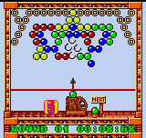 A colorful bubble-matching puzzle screen displays a grid of multicolored spheres clustered near the top, with a small launcher character positioned at the bottom center above a horizontal line. The play area is framed by a decorative orange and white pixelated border, with green grass-textured ground at the bottom and blue sky above. Red building structures appear on either side of the lower interface. The pixel art uses a vibrant palette of reds, blues, greens, yellows, and purples typical of Game Boy Color-era graphics.