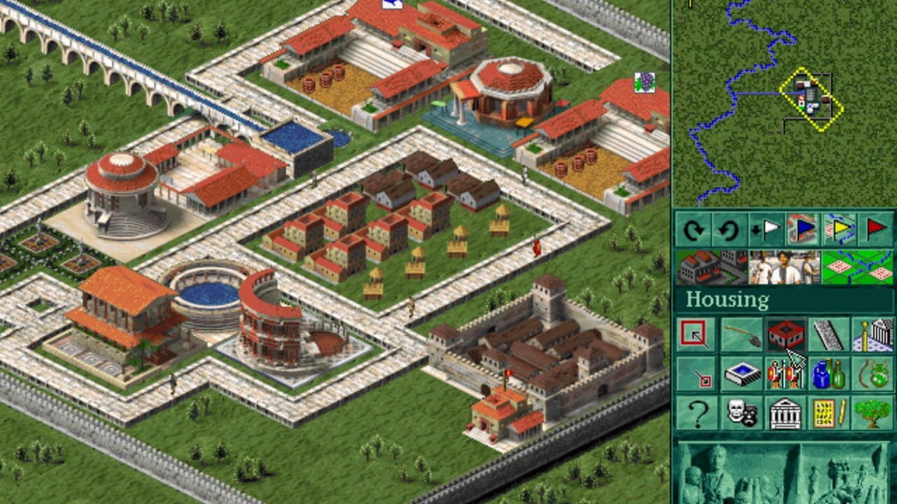 An isometric city-building view displays a Roman settlement with red-tiled buildings arranged in a grid pattern, including circular amphitheaters, a colosseum with blue interior, residential housing blocks with yellow details, and fortification walls. Green grass surrounds the stone pathways, with scattered trees in the borders. The right side shows a green UI panel labeled "Housing" containing various building and construction icons in a grid layout. A minimap in the top-right corner outlines the settlement with blue and yellow markers. The graphics use a low-resolution pixel art style typical of mid-1990s DOS games.