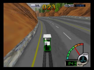 A green and white race car viewed from above drives down a desert highway with brown rocky terrain flanking both sides. Yellow center line markings divide the asphalt road. The HUD displays time remaining as 023 in the upper left, a speedometer in the lower right corner showing green and red zones, and a small map indicator in the bottom right. Rocky canyon walls and sandy dunes create a parallax effect on either side of the straightaway.