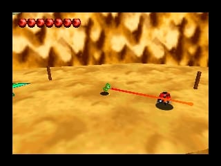 A red chameleon character stands on a sandy desert landscape with rocky outcrops and cliff faces in the background. Two small green insect-like enemies are visible on the terrain. The HUD displays six red circular health or life indicators in the top-left corner. The scene uses warm yellow and brown tones with 3D polygon graphics typical of N64 games, rendered with perspective depth showing the uneven, mountainous terrain.