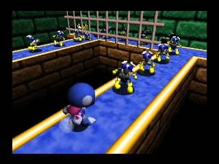 A blue chameleon character stands on a narrow blue platform with yellow edges in the foreground, facing a line of yellow and blue robot or enemy figures arranged in single-file formation extending into the distance. Green foliage forms a distant backdrop, with brown wooden structures and a blue fence visible along the platform edges. The scene uses a low polygon 3D art style typical of Nintendo 64 games, with flat colored surfaces and basic geometric shapes.