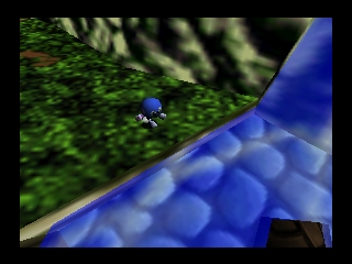 A small blue chameleon character stands on a curved blue surface in the foreground, with lush green foliage and terrain visible in the background. The 3D graphics show a colorful low-polygon environment typical of N64-era rendering, with a clear depth of field separating the character from the layered landscape behind it. The scene depicts an outdoor platforming level with natural vegetation and geometric blue platforms.