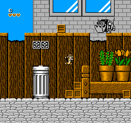 A side-scrolling level interior displays brown wooden walls with two windows at the top of a gray brick exterior wall. A white trash can sits on the left, while wooden crates, chairs, and potted plants with green foliage occupy the right side. Two cartoon mouse characters with simple dot eyes appear on the wooden surface—one in the upper portion and another lower down. The floor is rendered in light gray stone tiles, with a blue sky visible in the upper left corner showing yellow objects. The sprite art uses a limited color palette typical of NES-era graphics.