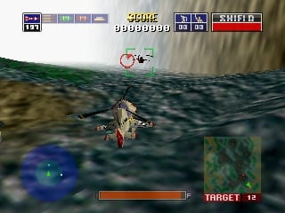 A helicopter viewed from above hovers in the center of the screen, facing upward over a landscape with green grass and gray rocky terrain. The HUD displays a red health bar at the bottom left, a compass or radar in the lower left corner showing blue and green markers, and a "TARGET: 12" indicator in red text at the bottom right. The top of the screen contains weapon and ammo counters, score display showing "0000000", and shield status in the upper right corner. The sky appears overcast with a flat, polygonal terrain aesthetic typical of early 3D console graphics.
