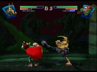 Two clay fighter characters face off in a fighting arena with a dark green and burgundy background. The left character wears red and holds a boxing stance, while the right character is tan-colored and mid-kick. Health bars for both fighters appear at the top of the screen in red and blue, with character portrait icons flanking them on either side. The arena floor is rendered in green with shadowy depth, and both fighters have a chunky, clay-textured 3D model appearance typical of N64-era graphics.
