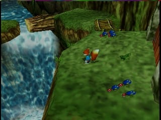 A green grass clearing with water and rocky cliffs visible on the left side. A brown squirrel character stands in the center-left area. Multiple blue and red numbered squares are scattered across the grass floor, likely representing collectibles or items. Wooden fence structures and logs are positioned around the perimeter of the area. The background shows brown rocky formations and green vegetation typical of a forest environment rendered in low-polygon 3D graphics.