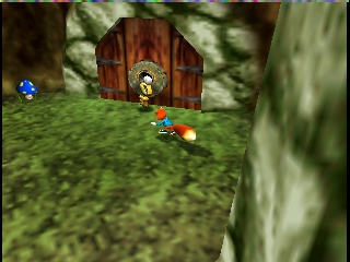 A character stands in a grassy outdoor area with a large wooden barn door ahead. On the right side, a thick tree trunk frames the scene. The character holds a weapon and carries supplies on their back. The upper left corner displays a small blue HUD icon. The environment uses 3D polygonal rendering with detailed textures showing grass, wood, and foliage. The camera is positioned at ground level, creating a first-person perspective view of the landscape.
