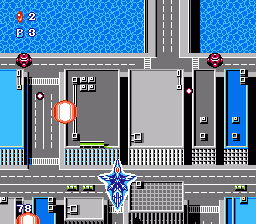 A vertical scrolling shooter displays an urban cityscape with gray and blue buildings arranged along a street. A blue player aircraft with a distinctive star emblem flies near the bottom center of the screen. Red enemy ships appear in the upper left corner. The HUD shows a score of 0 and 0 in the top left, with additional numerical displays along the top edge. A yellow fence line separates the playfield from the street below, and the overall 8-bit sprite-based visual style uses a limited palette of blues, grays, reds, and whites against a light blue scrolling background.