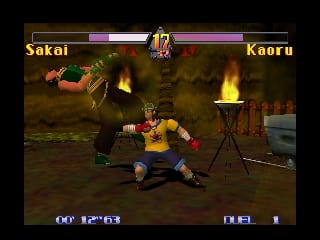 Two fighters engage in combat on a brown arena floor with fire and wooden structures visible in the background. The character on the left wears green clothing while the opponent on the right wears yellow and red. Health bars labeled 'Sakai' and 'Kaoru' appear at the top in purple and pink, with the match timer displaying '12' in the center. The lower left shows 'GO JUSTICE' text while the lower right displays 'DUEL'. The 3D graphics use earthy tones with volumetric fire effects, characteristic of Nintendo 64-era polygon rendering.