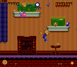 A side-scrolling platformer level displayed in SNES-era pixel graphics with a warm brown wooden interior theme. The blue-clad protagonist stands on the main floor amid furniture including a dark dresser and table. Two elevated wooden platforms with green grass patches occupy the upper portion, one containing a pink character or enemy. The HUD spans the bottom with a yellow coin counter on the left, health/lives display in the center, and orange circular elements on the right. Vertical wooden wall panels form the background, creating a domestic indoor setting with distinct vertical striping.