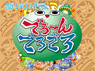 A large green frog character with wide eyes and an open mouth dominates the center of the title screen. Japanese text appears at the top in blue and yellow, with the game's stylized logo displayed prominently in green and blue below the frog's head. Small colorful game elements and icons are scattered across the bottom portion of the screen against a tan patterned background. Decorative ornamental designs frame the upper corners in gold and brown tones.