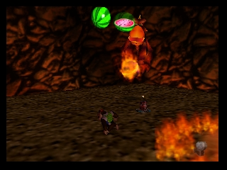 A dark, textured ground environment with a small character figure positioned in the lower-left area. Two large colorful projectiles—one bright green and one orange-red—descend from the top center of the screen. Flames and fire effects glow orange in the lower-right corner. The background shows brown, cracked terrain textures. The 3D perspective and low-polygon geometry are characteristic of Nintendo 64-era graphics.