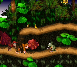 Two anthropomorphic apes stand on a wooden log suspended over a jungle backdrop, flanked by glowing green foliage and vines. The characters wear red and brown clothing, positioned in the center-left and right portions of the screen. Thick wooden branches form the main platform, with layered vegetation in dark greens and reds creating depth behind them. Pixelated 16-bit sprite graphics with a warm color palette dominate the scene, typical of SNES-era platformer visuals.
