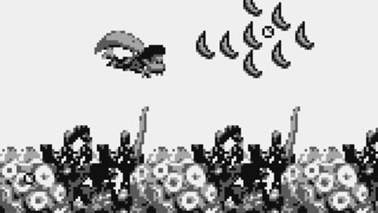 A Game Boy screenshot showing a helicopter with a pilot in the upper left, facing right toward a cluster of banana sprites arranged in the upper right. Below stretches a repeating landscape of pixelated terrain with industrial structures, smokestacks, and white circular objects resembling factory elements or pipes. The monochrome palette uses black, white, and gray pixels typical of the Game Boy hardware.