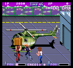 A military helicopter with yellow-green fuselage and brown cargo pod hovers above a gray concrete surface, with three player characters in fighting stances positioned below it. The arcade UI displays a score counter at bottom left, health bars for two players at the bottom corners, and a purple header bar at top showing round information and remaining time. The sprite art uses bright primary colors against a neutral background in a 1988 arcade resolution style.