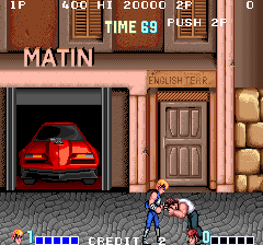 A side-scrolling beat-em-up scene displays two player characters in blue martial arts outfits positioned in the center-lower portion of the screen, facing a red car visible through an open garage door on the left. The background shows a brick building facade with wooden doors labeled 'HONGH TERA' and scattered barrels. The top HUD displays score values (1F, 266, 14, 26000, 2F), time counter at 69 seconds, and a PUSH indicator. Pixel art sprites and brown-toned environment colors are consistent with arcade-era 16-bit graphics.