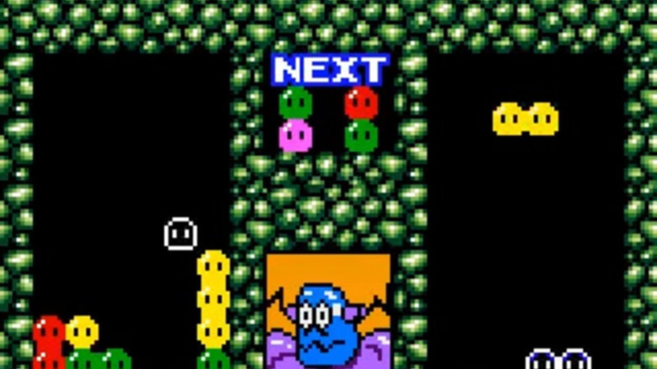 A puzzle game board displays a blue character in the center surrounded by falling colored beans. A blue "NEXT" label at the top shows upcoming pieces: green, red, pink, and green beans. The playfield is bordered by green brick walls on both sides. Yellow bean chains occupy the left edge, while two yellow beans appear in the upper right. Small colored enemies (red, yellow, green) populate the left and upper edges. The black background contrasts with the pixelated 8-bit sprite art typical of early handheld gaming.