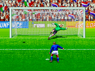 A soccer match in progress on a bright green field with a goalkeeper in green positioned at the center goal. A player in blue uniform stands in the foreground holding the ball near the penalty area. The goal net is clearly visible with yellow crossbar and white netting. A colorful crowd of spectators fills the stadium background with red, white, and blue clothing. Advertisement boards line the top perimeter of the field. The sprite-based graphics use bright, solid colors typical of early 1990s arcade hardware.