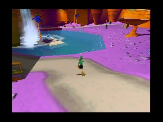 A small character avatar stands in the center of a sand-colored arena floor, surrounded by bright magenta and cyan-colored terrain platforms and structures. A fountain or water feature with white spray is visible in the background to the left. The scene uses a vibrant pastel color palette typical of Nintendo 64-era 3D graphics, with solid-colored geometric forms defining the play space. The camera angle shows a third-person overhead perspective of the game environment.