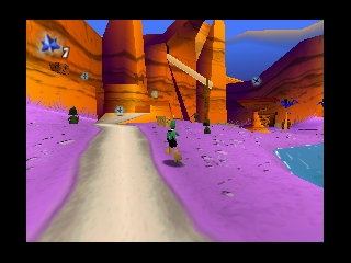 A first-person perspective view down a white road stretches toward colorful rock formations and orange pillars in the distance. The landscape is rendered in warm orange and blue tones with a bright pink/magenta ground plane on either side of the path. Small purple objects are scattered across the terrain, and the sky displays a gradient from orange to blue. The 3D environment uses flat-shaded polygonal geometry typical of early N64 games, with minimal texture detail and bold color blocking creating a stylized desert or alien landscape setting.