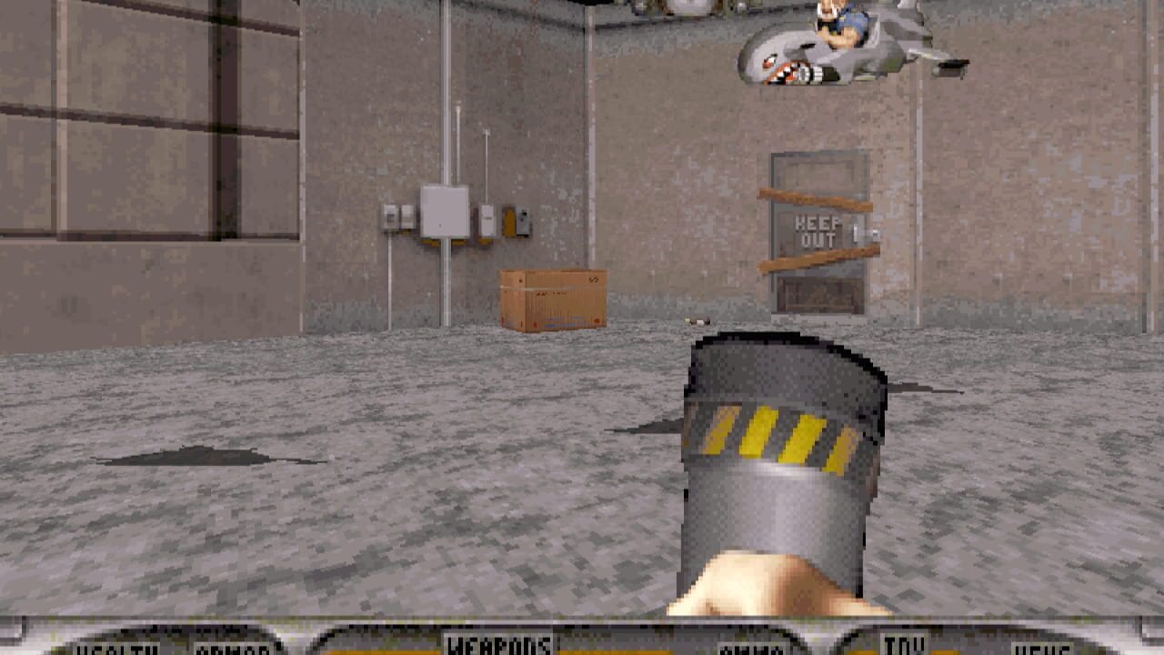 A first-person view of an industrial warehouse interior with concrete walls and flooring. The player's weapon—a gray cylindrical device with yellow and black hazard stripes—occupies the bottom right corner. The scene contains wooden crates, electrical boxes on the walls, wooden shelves marked "HERE OUT", and various gray structural elements. An enemy character appears in the upper right corner of the room. The HUD displays health, armor, and weapons information at the bottom of the screen in a yellow-text interface typical of early 1990s DOS games.