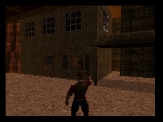 A character in dark clothing stands in a sandy courtyard between two wooden buildings with boarded windows. The low-polygonal 3D environment uses a muted brown and tan color palette typical of early N64 graphics. The buildings feature multiple window panes and weathered wooden textures. The ground is rendered as coarse sand or dirt with visible pixelation and texture mapping consistent with the console's technical limitations. Shadows cast from the structures indicate a light source from the upper right.