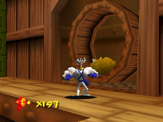 Earthworm Jim stands on a wooden platform in the center of the frame, wearing his signature blue suit and helmet. Behind him is a large circular wooden tunnel entrance set into wooden walls with vertical paneling. A yellow flower or plant protrudes from the tunnel opening. The lower-left corner displays a red health or lives indicator with the number 3 and Japanese characters. The scene uses warm brown wood tones with green foliage visible at the top edge, rendered in 3D polygon graphics typical of late-1990s Nintendo 64 games.