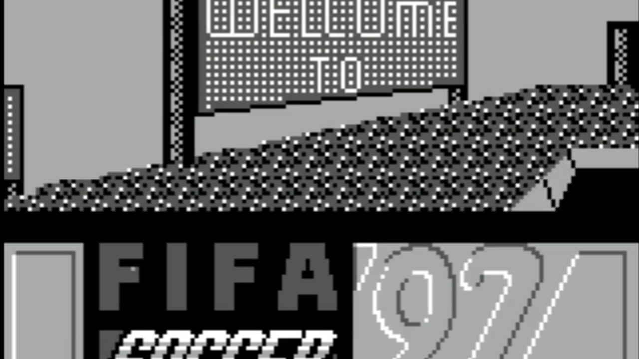 A monochrome Game Boy screenshot displays a pixelated soccer stadium view from an overhead perspective. A dotted pattern represents the grass field with a checkered border fence visible at the top. A pixelated building structure with white dots appears in the upper portion of the screen, suggesting a stadium grandstand. At the bottom, large gray block letters spell 'FIFA' with partial white text '97' visible to the right, indicating the game title. The scene uses the characteristic low resolution and limited color palette typical of Game Boy sports games from the 1990s.